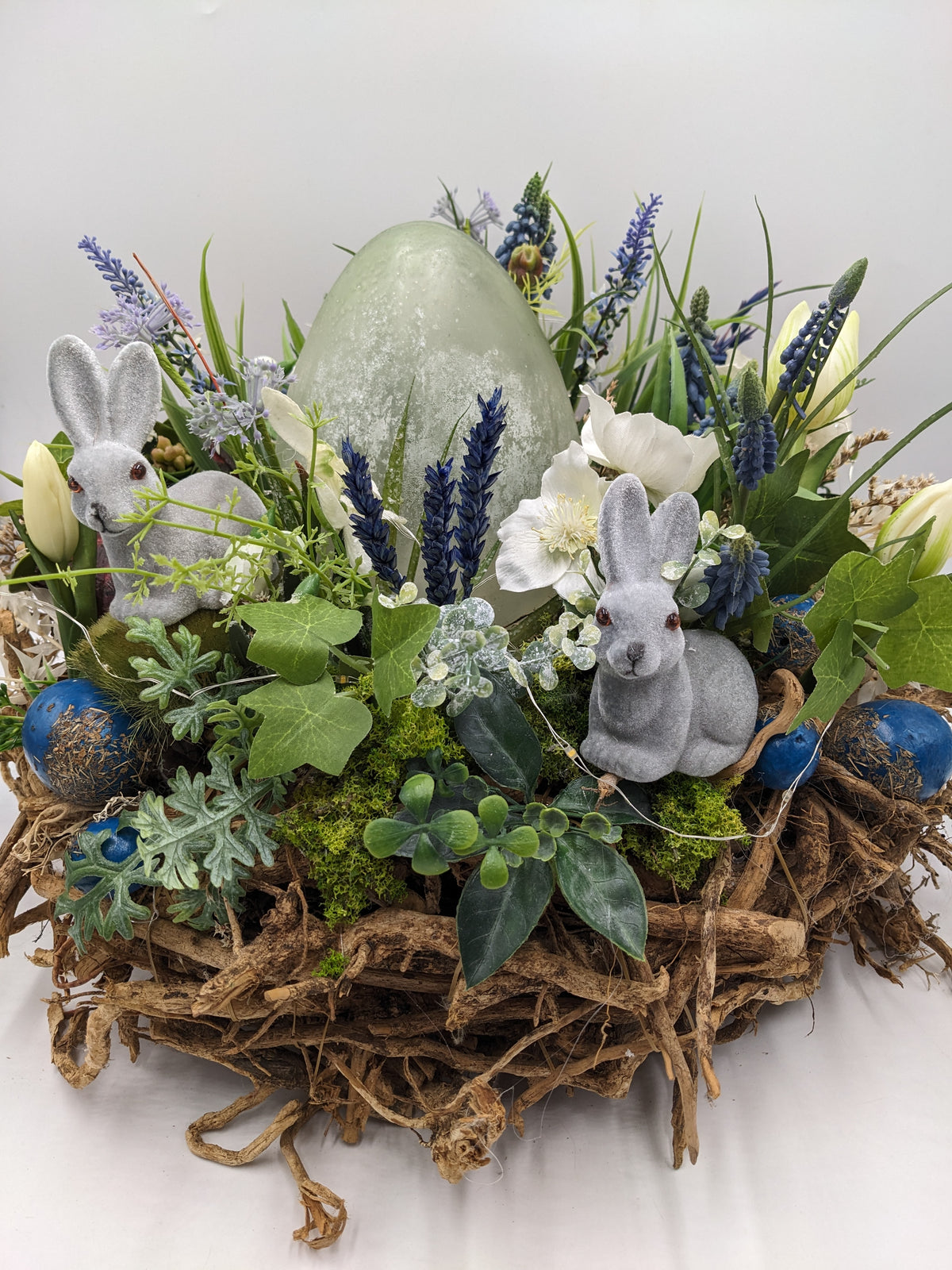Ostergesteck mit grünem Osterei, zwei grauen Samthasen, blauen Hyazinthen und weißen Blüten in Wurzelschale