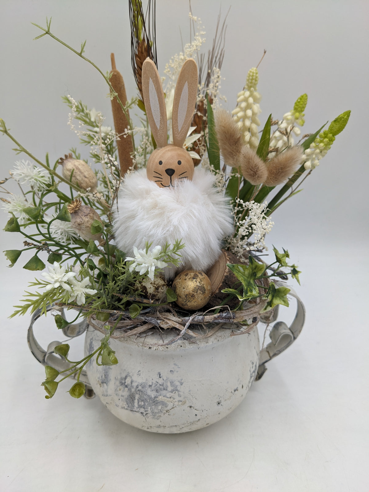 Frühlingsgesteck in Creme und Weiß mit Hasenfigur, Efeu, Mohnkapseln und Wachtelei, Nahansicht von vorn