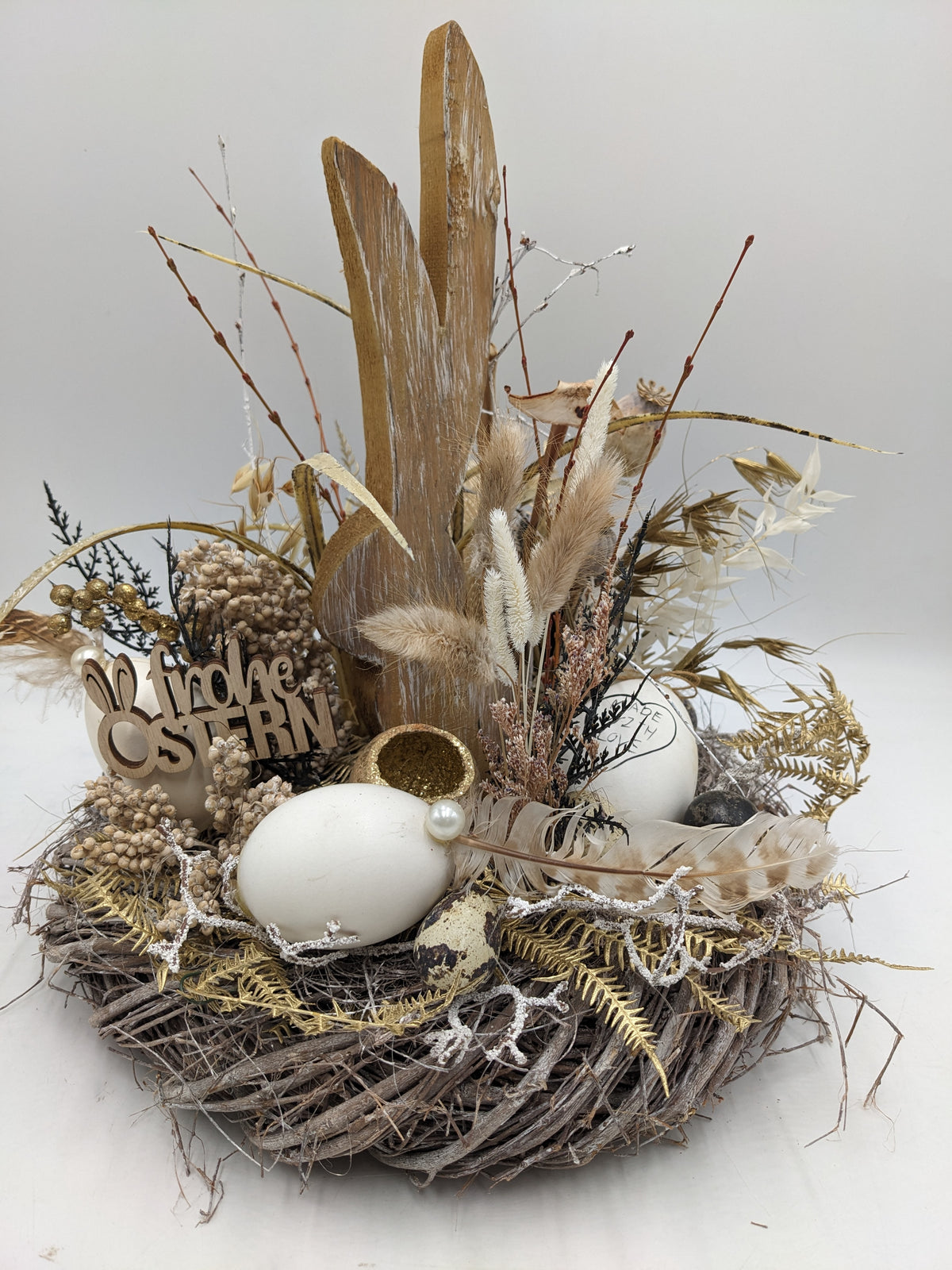 Ostergesteck von der Seite: Holzhase, Schriftzug Frohe Ostern, goldene Eierschale und Wachtelei im Nest