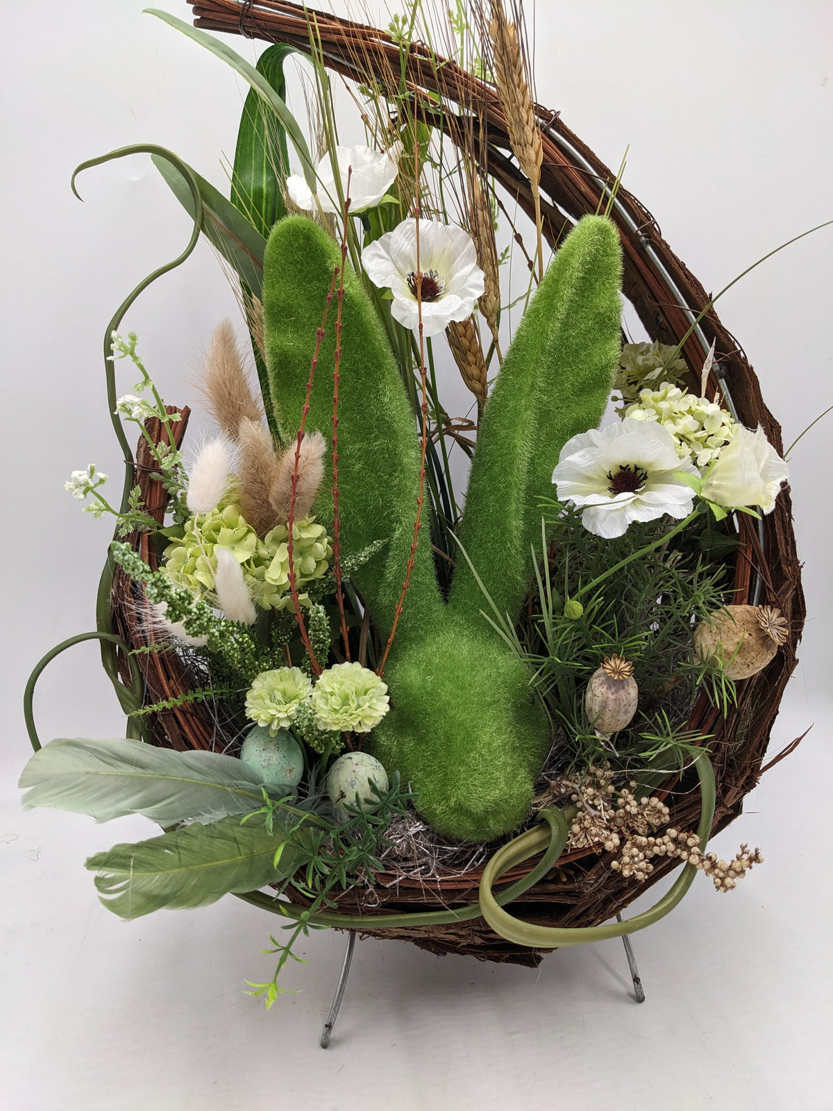 Ostergesteck von vorn: grüner Hase im Nest mit mintfarbenen Eiern, grünen Nelken und weißen Blüten
