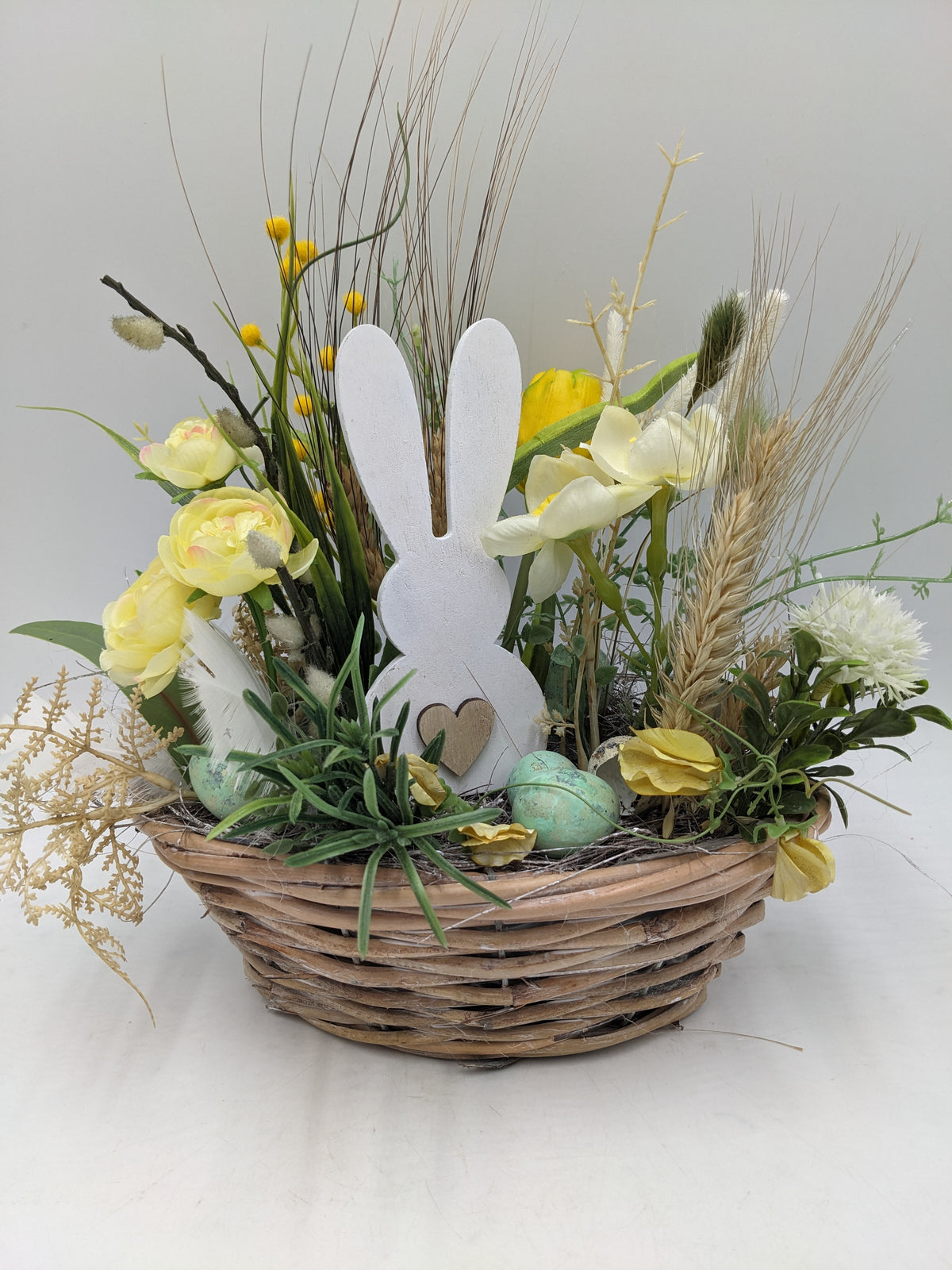 Ostergesteck in Weidenschale mit weißem Holzhasen, gelben Ranunkeln, Tulpen, Gräsern und Ähren