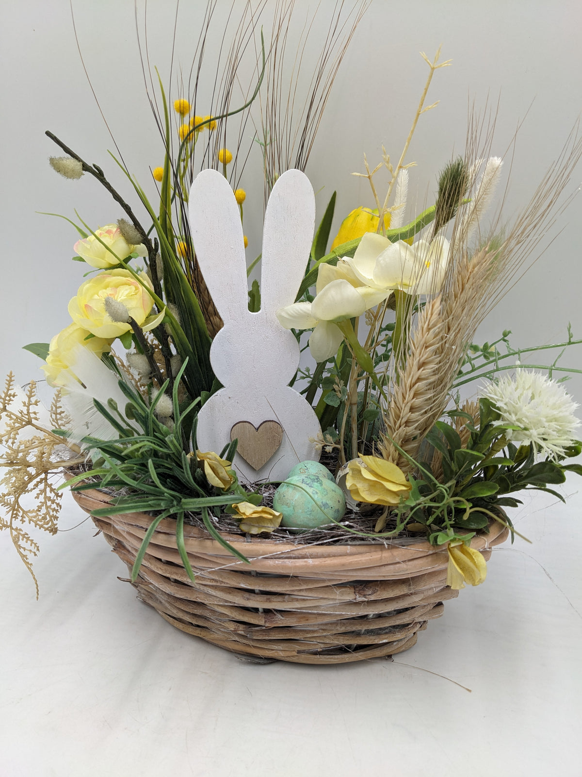 Osterkorb von vorn: weißer Hase mit Herz, türkisgrünes Ei, gelbe Blüten und Weidenkätzchen im Nest
