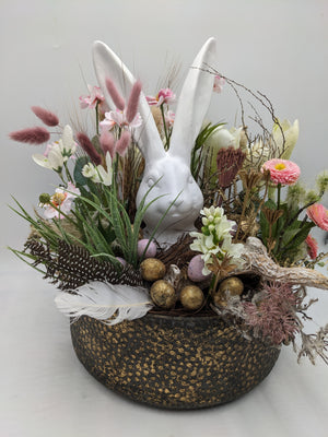 Frühlingsgesteck von der Seite: Hasenfigur, Gänseblümchen, Trockengräser und Ostereier in runder Schale