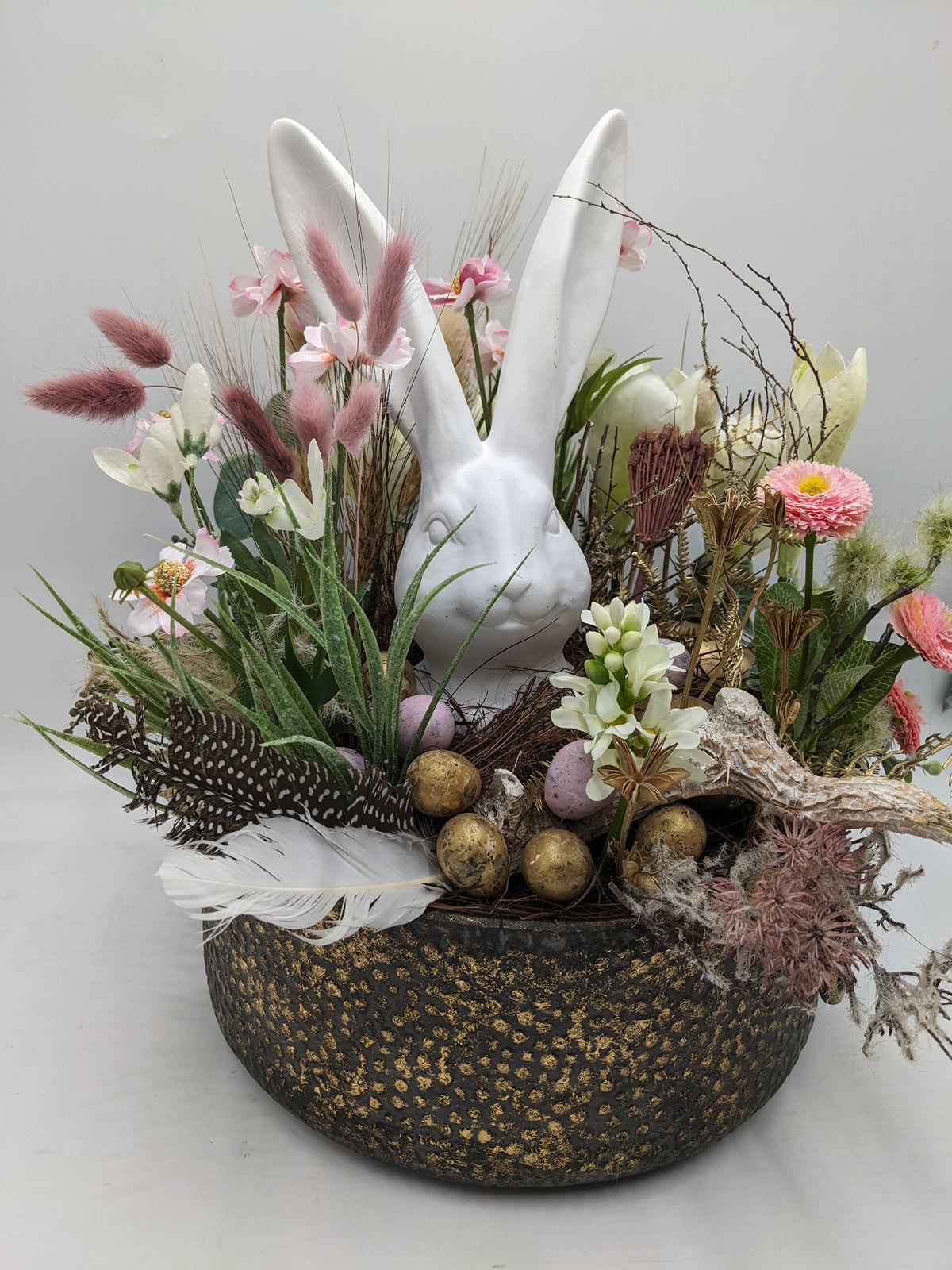Frühlingsgesteck von der Seite: Hasenfigur, Gänseblümchen, Trockengräser und Ostereier in runder Schale
