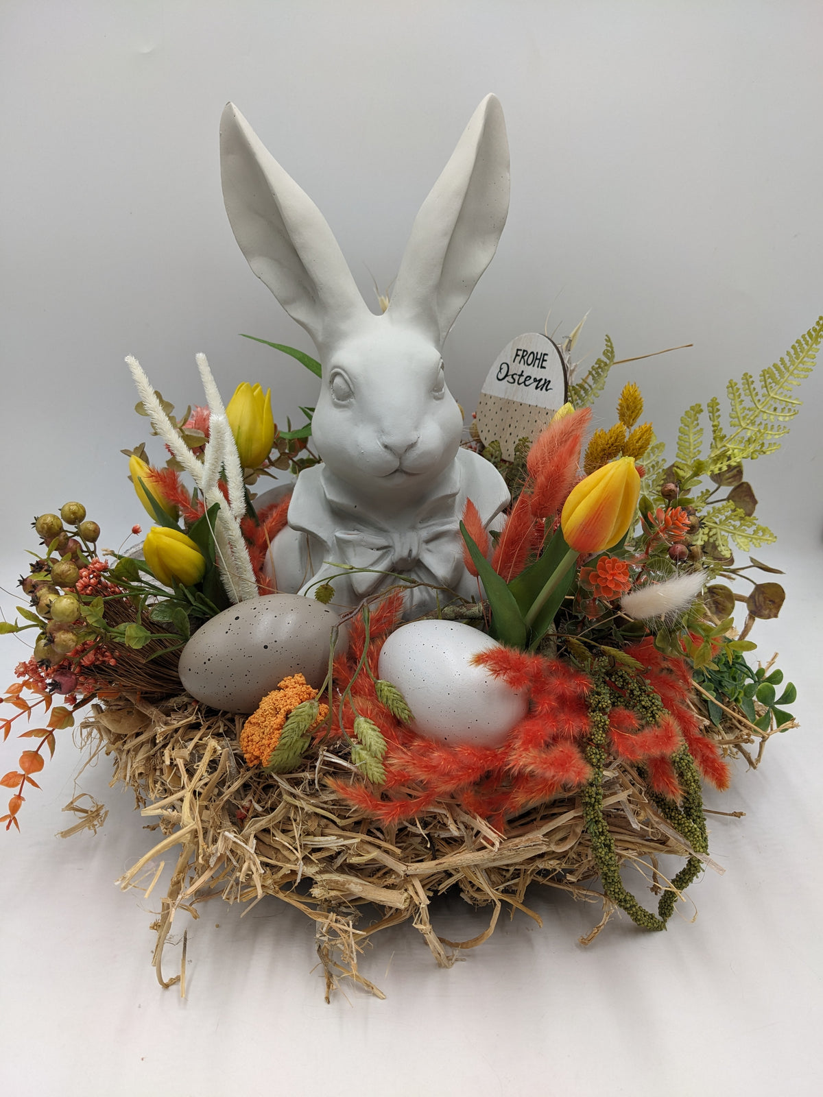 Ostergesteck im Strohnest mit weißer Hasenbüste, gelben Tulpen, orangen Gräsern und zwei gesprenkelten Eiern