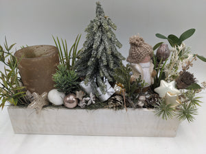 Wintergesteck in weißer Holzschale mit verschneitem Tannenbaum, Wichtelfigur, Zapfen und braunem Teelichtglas