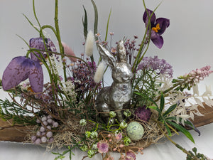 Nahaufnahme: silberner Hase im Nest aus Holzwolle, grünes geflecktes Ei, lila Beeren und violette Krokusblüte