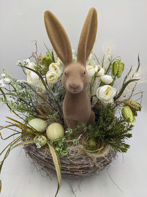 Frühlingsgesteck von vorn: Hasenfigur zwischen weißen Blüten, grünen Zweigen und gesprenkelten Ostereiern im Nest