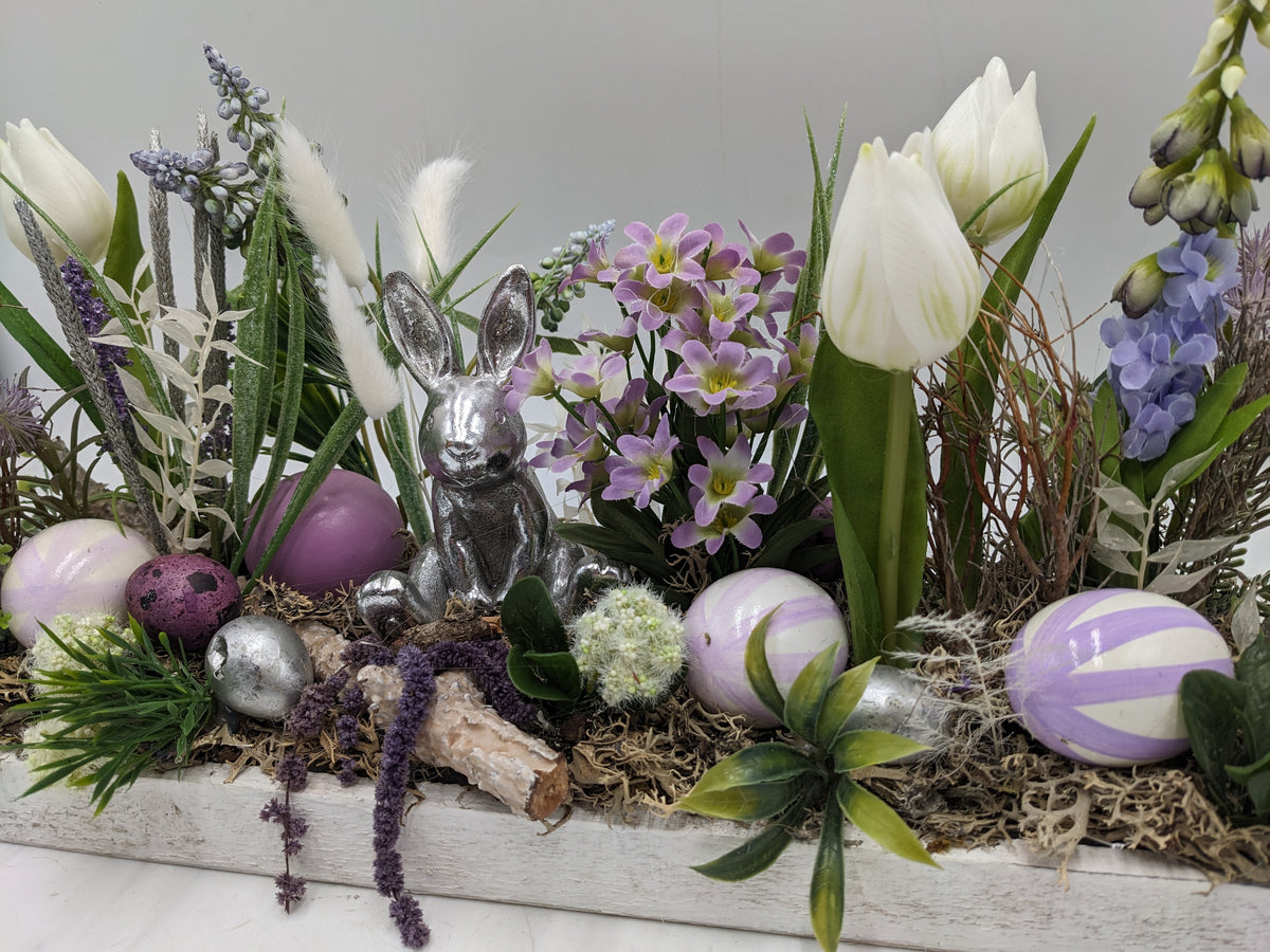 Nahaufnahme: silberner Hase zwischen lila-weiß gestreiften Ostereiern, Moos, Gräsern und weißen Tulpen