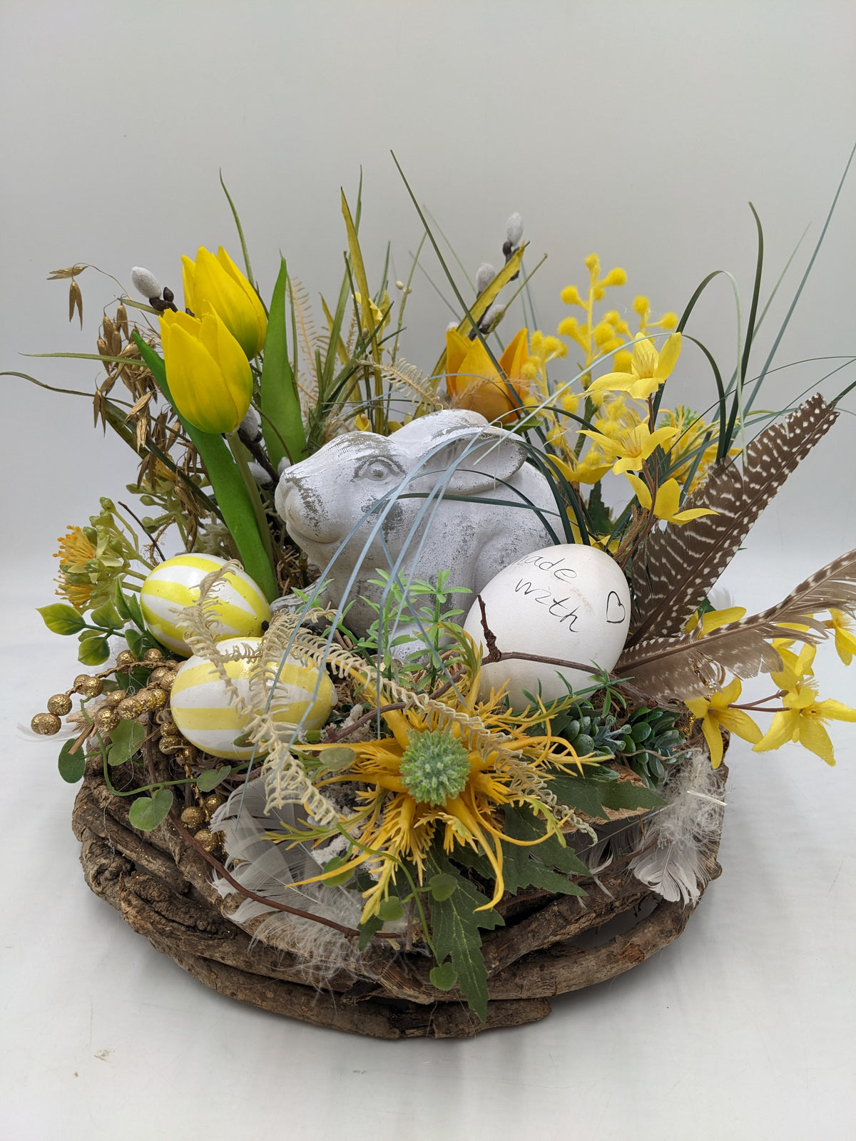 Ostergesteck mit weißem Hasen, gelben Tulpen, gestreiften Eiern und Federn auf rundem Rindenkranz