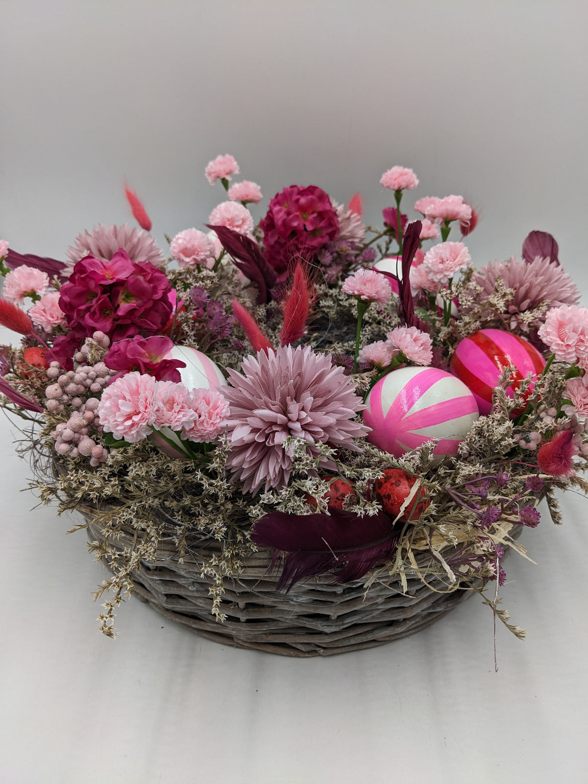 Osterkranz im grauen Flechtkorb mit rosa Nelken, Fuchsia-Blüten, pink-weiß gestreiften Eiern und Trockengräsern
