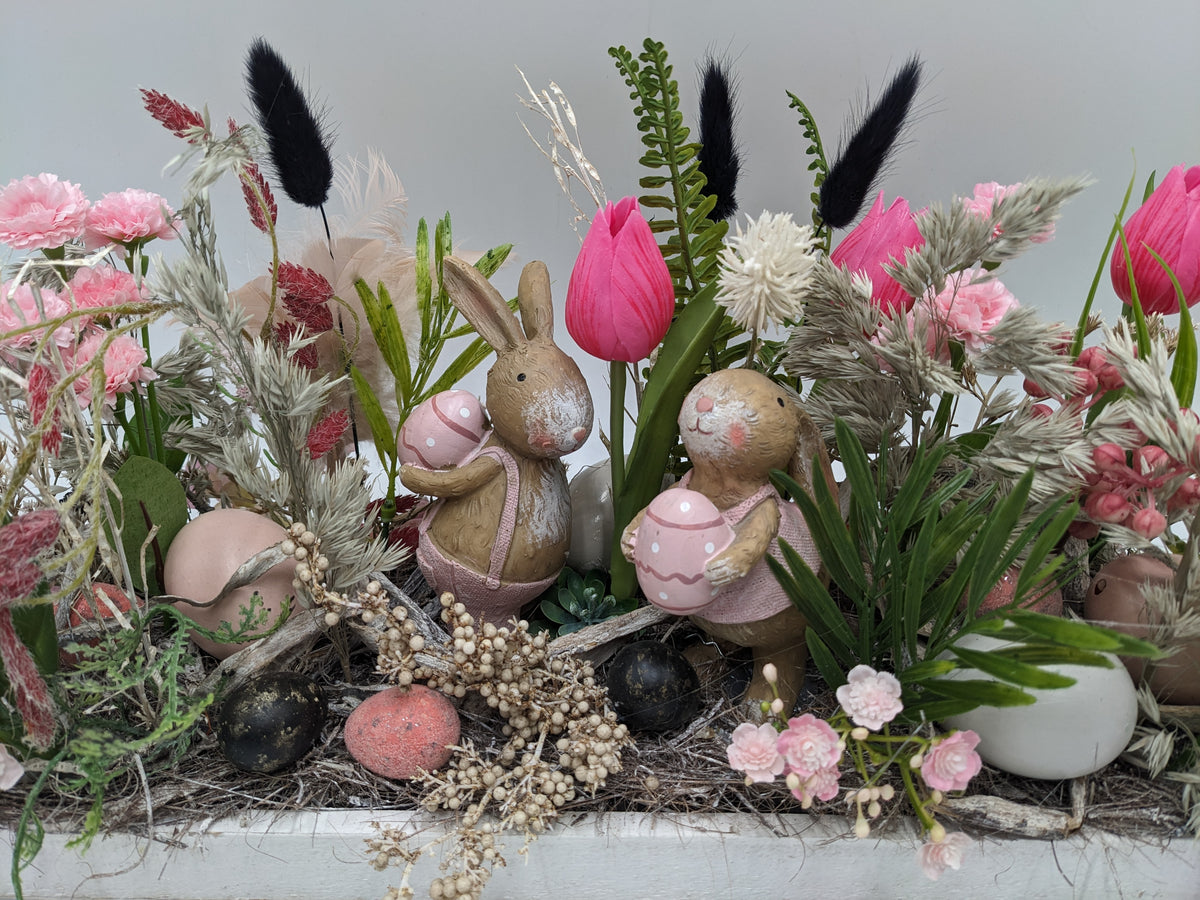 Nahaufnahme der beiden Keramikhasen mit rosa gepunkteten Eiern zwischen Tulpen, Nelken und Moos