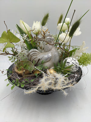 Ostergesteck mit silberner Hühnerfigur, weißen Tulpen, Gräsern und goldenen Eiern in schwarzer Schale