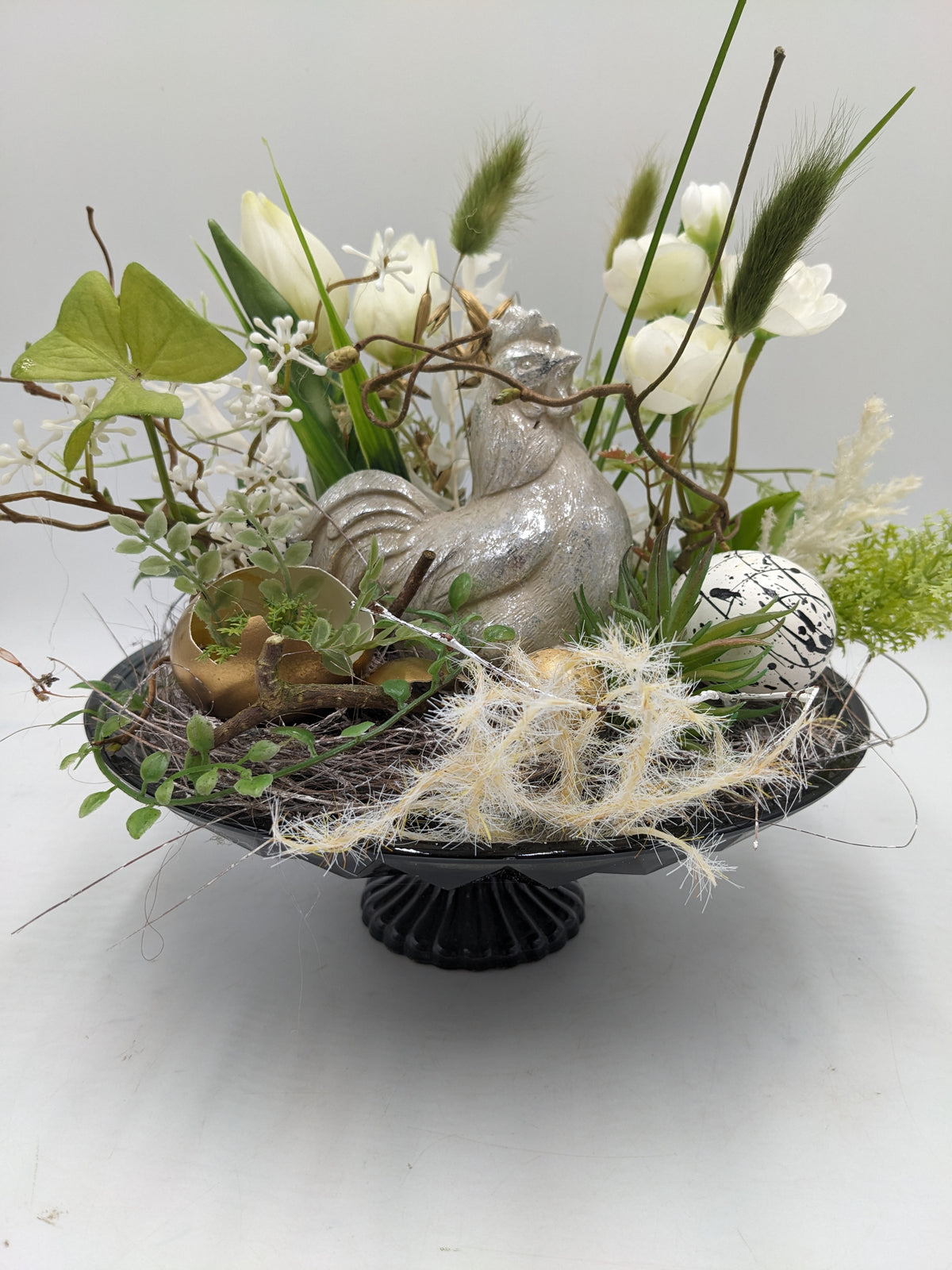Frühlingsgesteck auf schwarzer Fußschale mit Huhn, Nest, gesprenkeltem Ei und weißen Blüten, seitlich