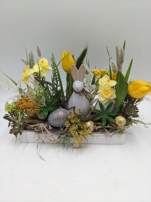 Frühlingsgesteck von vorn: Keramikhase zwischen gelben Tulpen, Narzissen, Gräsern und goldfarbenen Ostereiern