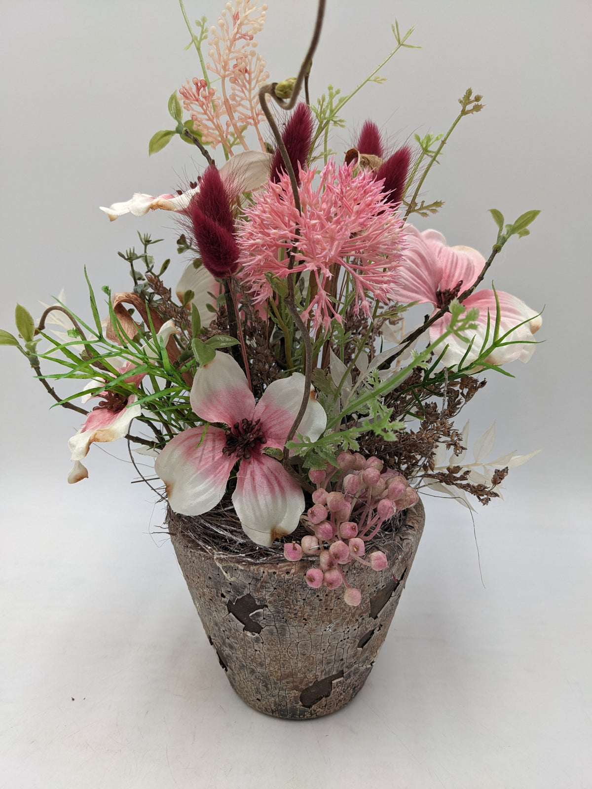 Frühlingsgesteck mit rosa-weißen Blüten, pinkfarbener Distel und dunkelroten Gräsern in rustikalem Topf