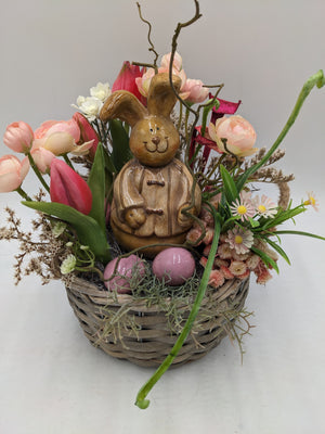 Frühlingsgesteck von vorn: brauner Keramikhase im Weidenkorb, umgeben von pinken Tulpen, rosa Ranunkeln und Trockengräsern