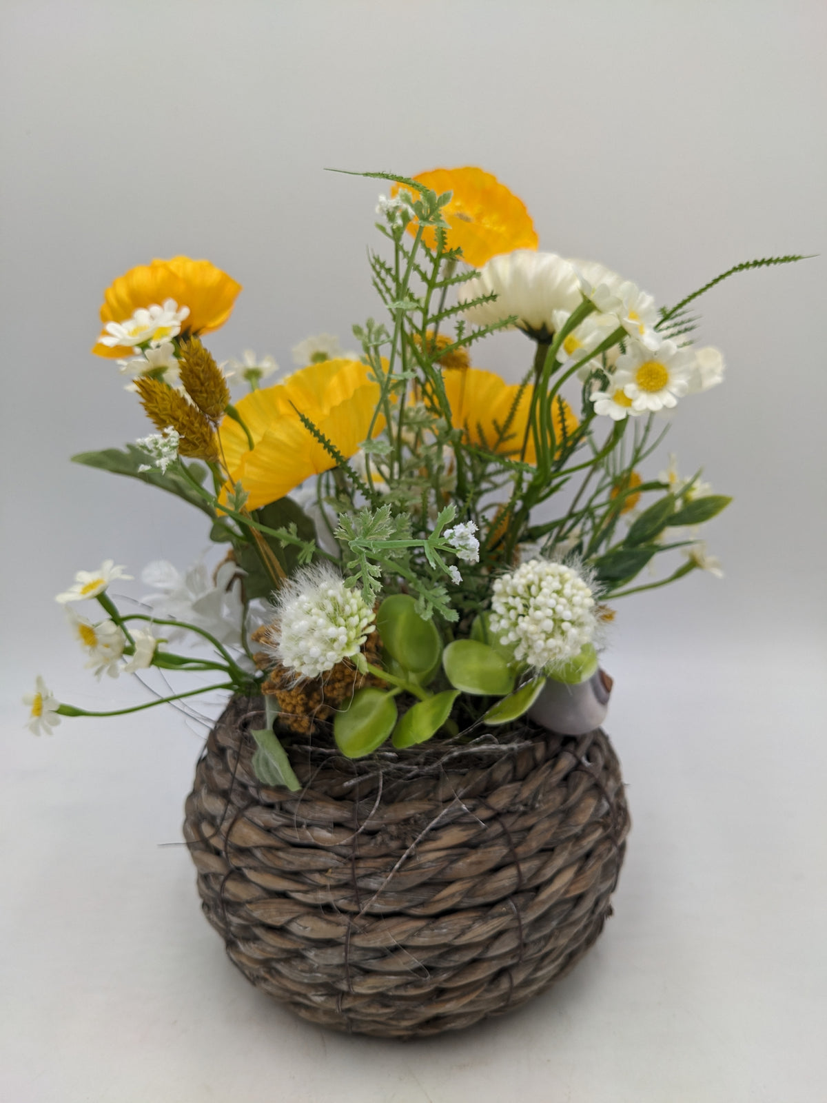Frühlingsgesteck mit gelben Mohnblüten, weißer Kamille und Gräsern in rundem Flechtkorb, von oben gesehen