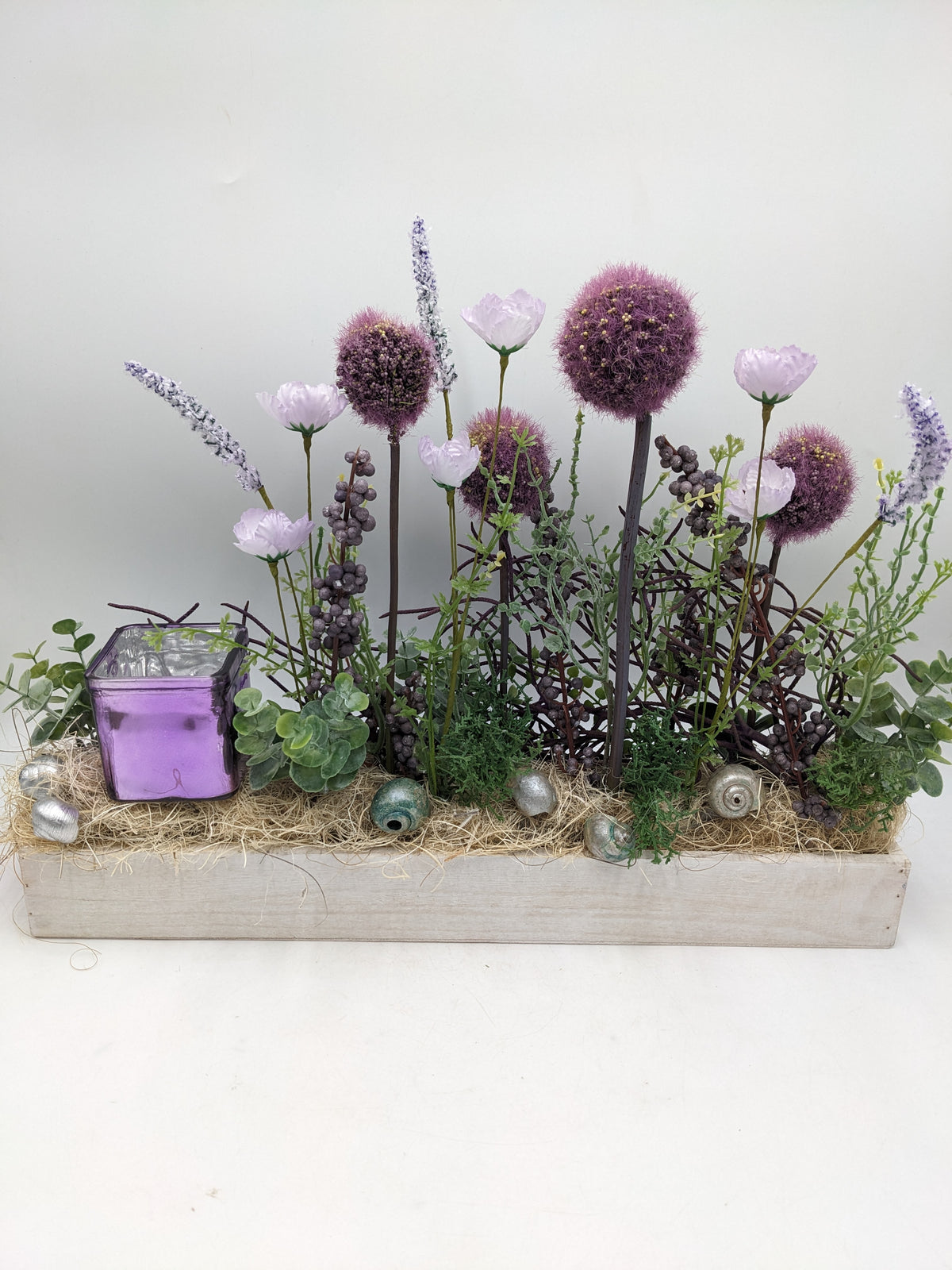 Längliches Frühlingsgesteck in weißer Holzkiste mit lila Kugelblüten, Mohnblüten und violettem Glaswindlicht