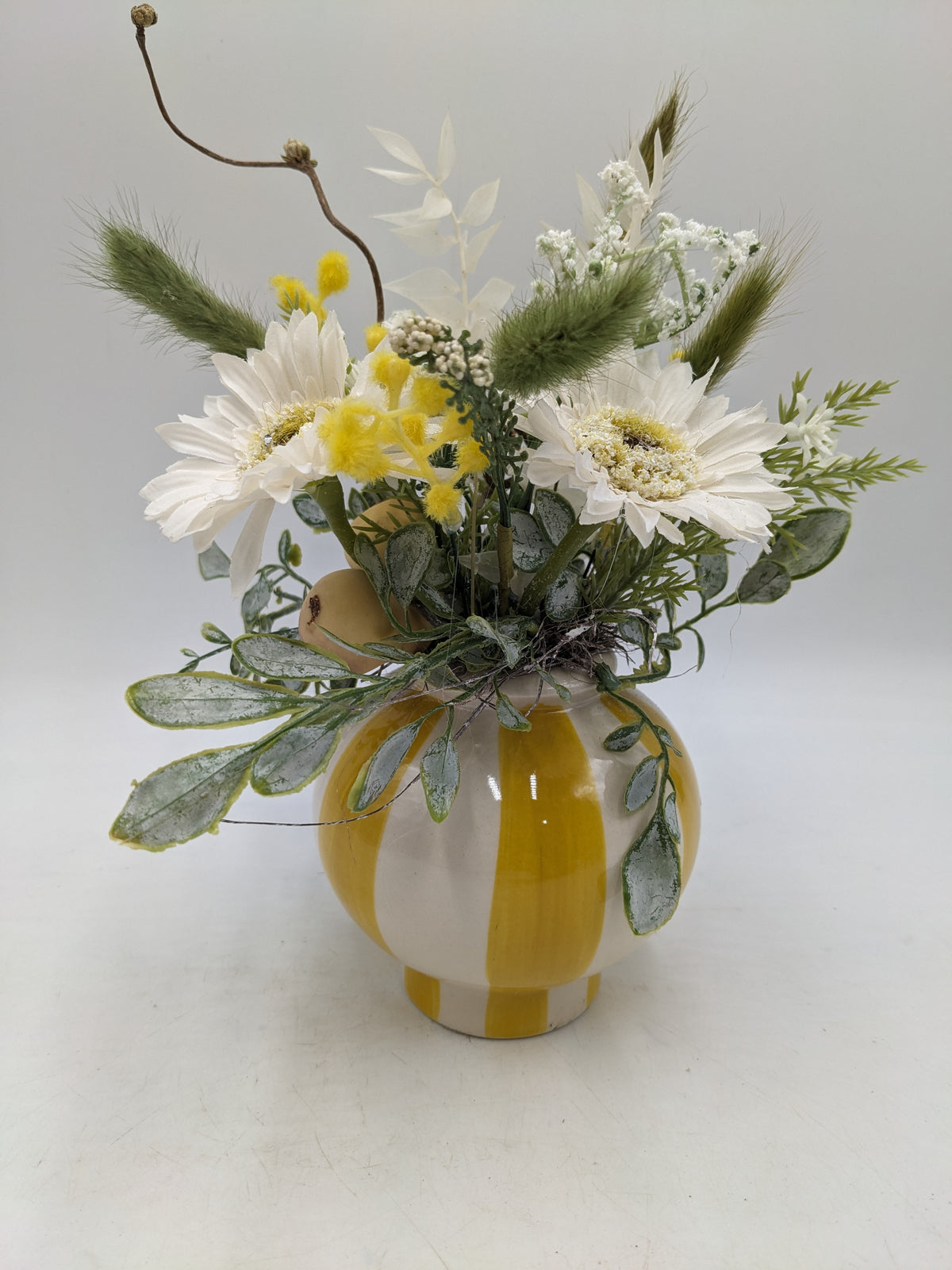 Frühlingsgesteck mit weißen Margeriten, gelben Mimosen und grünen Gräsern in gelb-weiß gestreifter Keramikvase