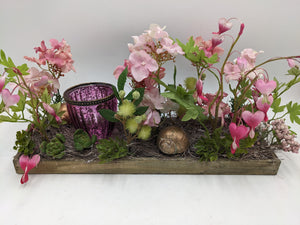 Langes Frühlingsgesteck in Rosa mit Tränendem Herz, Hortensien und purpurfarbenem Windlichtglas auf Holzschale