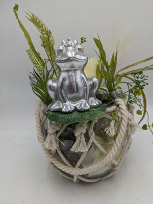 Sommergesteck mit silbernem Frosch auf Seerosenblatt über Glasgefäß im Kordelnetz, mit Gräsern und weißer Blüte