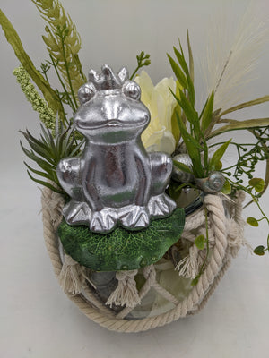 Nahaufnahme des silbernen Froschs mit Krone, daneben cremeweiße Blüte, grüne Ranken und silberne Schneckenhäuser
