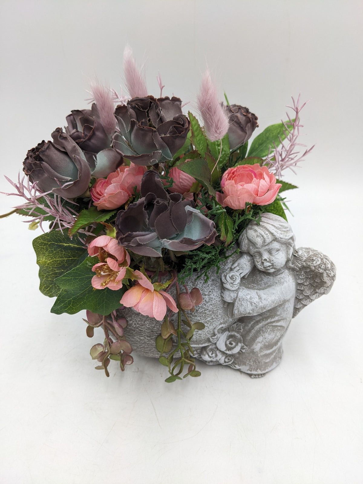 Trauergesteck mit mauvefarbenen Rosen, rosa Blüten und Gräsern in grauem Gefäß mit schlafender Engelsfigur