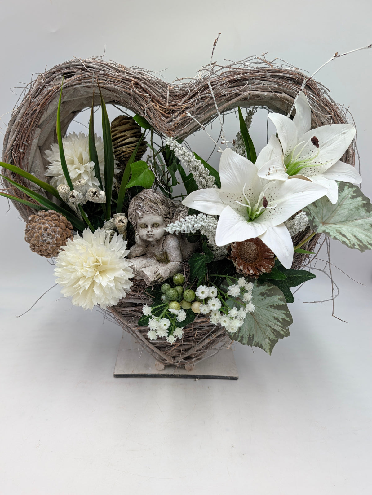 Herzförmiges Trauergesteck aus hellem Geflecht mit weißen Lilien, cremefarbenen Dahlien, Efeu und Engelsfigur auf Holzfuß