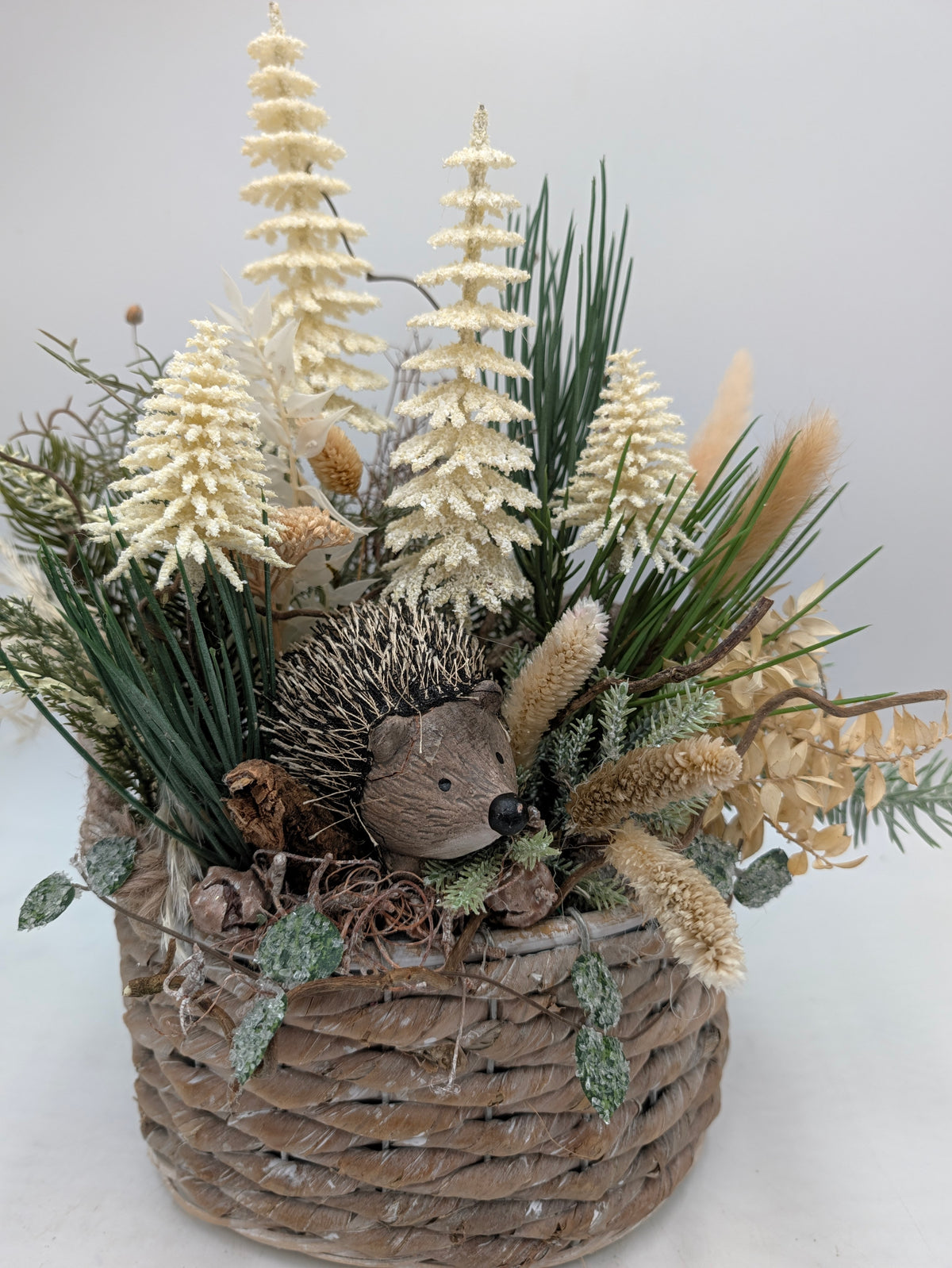 Nahaufnahme des Wintergestecks: Igel zwischen cremefarbenen Tannen, Gräsern und bereiften Blättern am Korbrand