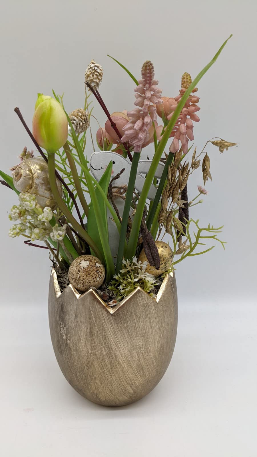 Ostergesteck im goldenen Eiertopf mit rosa Traubenhyazinthen, gelber Tulpe, Holzhase, Wachtelei und Schneckenhaus