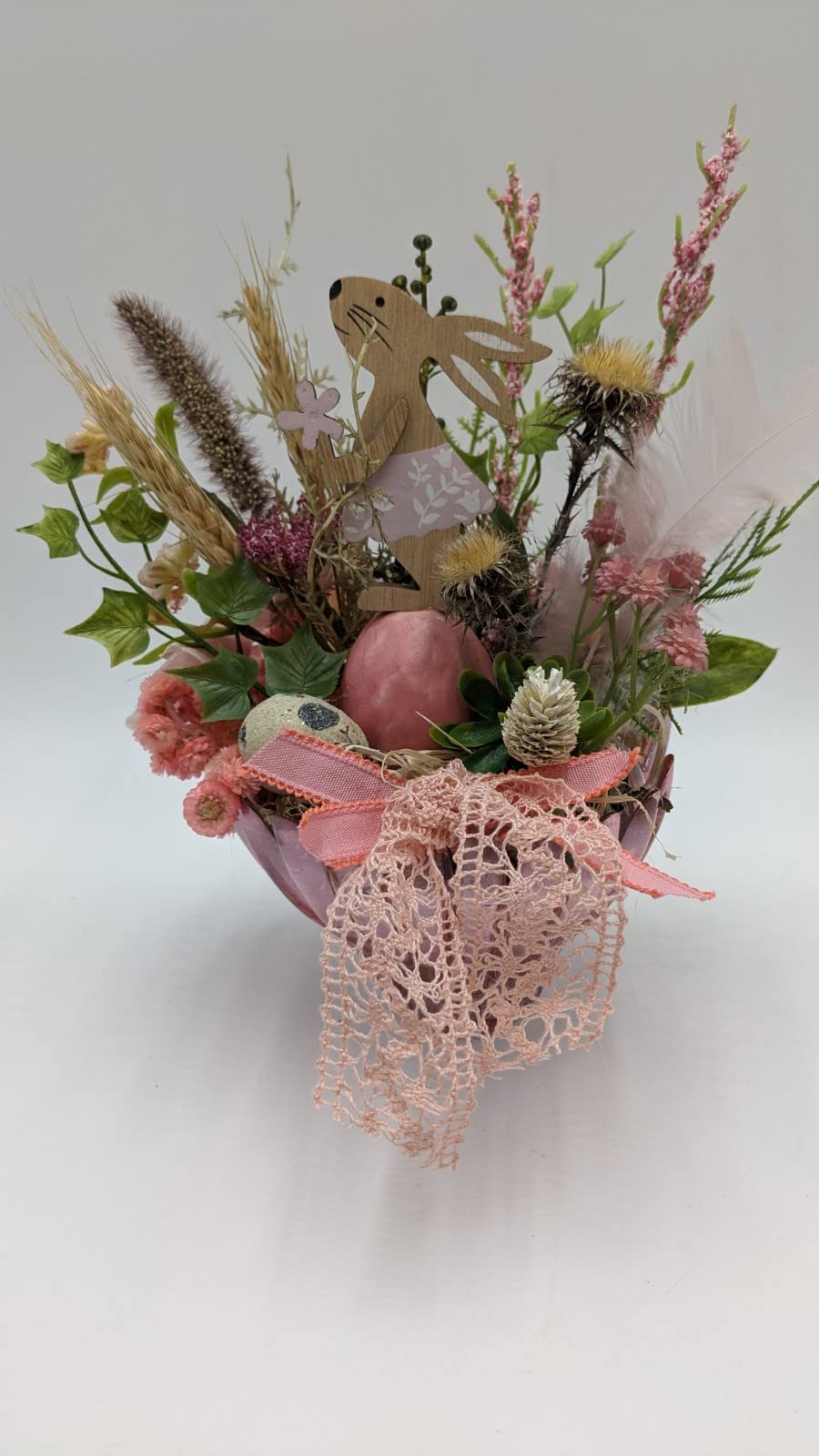 Ostergesteck mit Holzhase, rosa Ei, Gräsern, Federn und Spitzenband in rosa Glasschale, Nahaufnahme