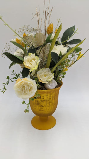 Tischgesteck mit cremefarbenen Rosen, Gräsern und Schmetterling in gelbem Pokal mit Fuß, seitlich fotografiert