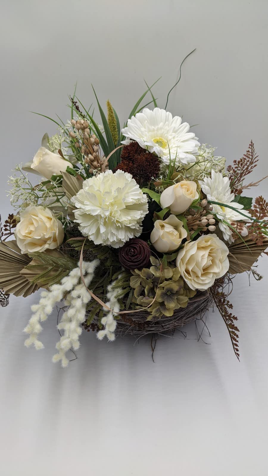 Tischgesteck von oben: weiße Gerbera, Nelke, Rosenknospen, dunkelrote Ranunkel und helle Palmblätter im Weidenkorb