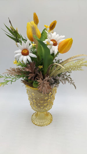 Seitenansicht des Frühlingsgestecks mit orangefarbener Tulpe, Gräsern und Zapfen über dem gelben Glasfuß