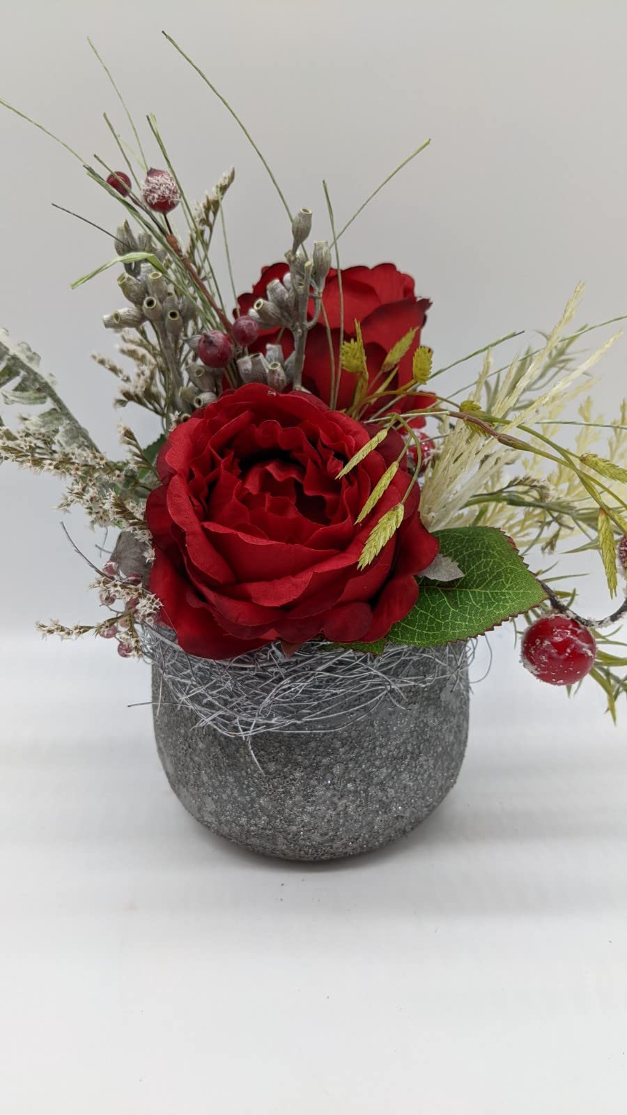 Tischgesteck mit zwei roten Rosen, bereiften Beeren und hellen Gräsern in grauem Topf mit körniger Oberfläche