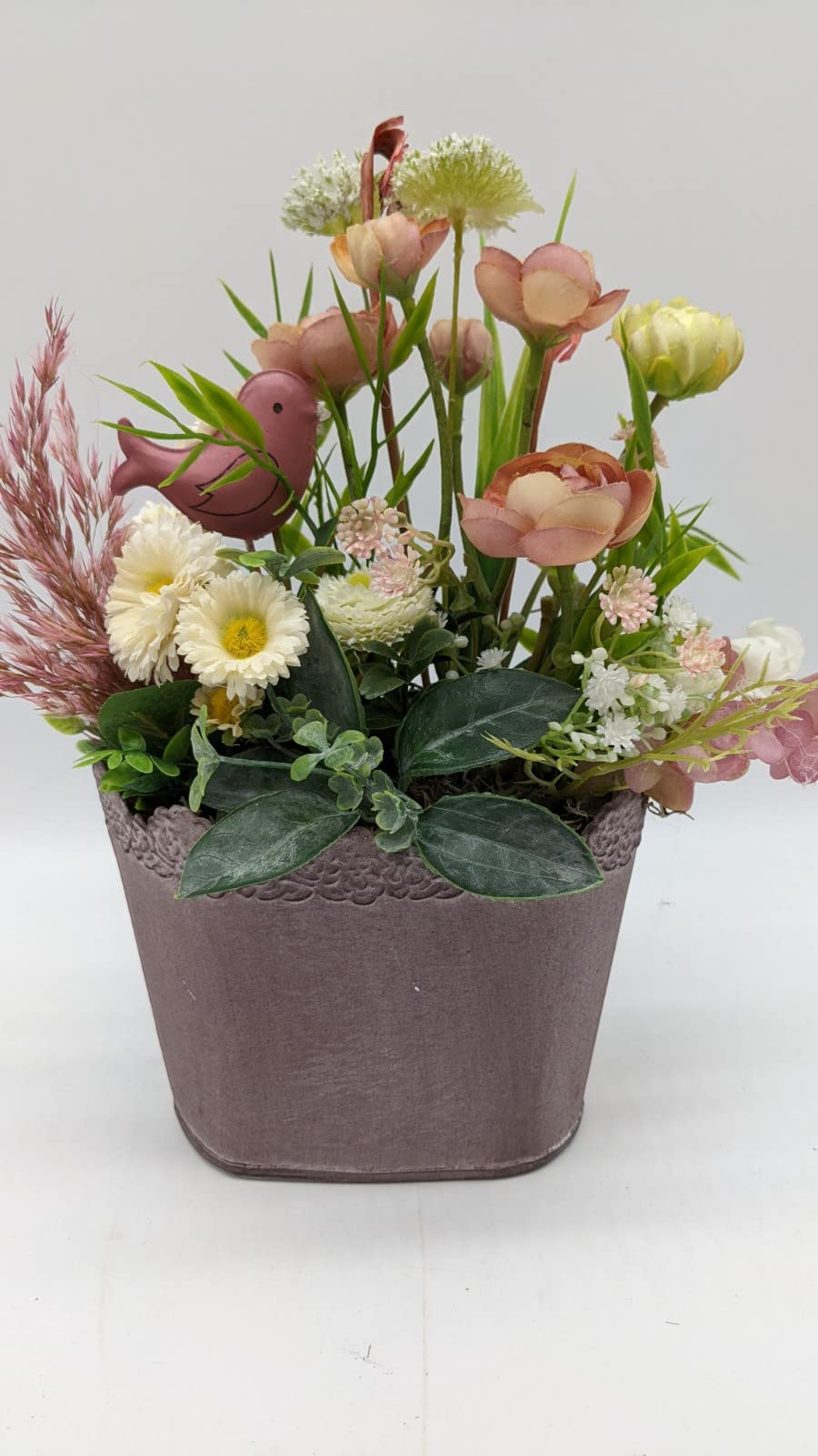 Tischgesteck in rosa Topf mit Ranunkeln, cremefarbenen Bellis, Gräsern und kleinem rosa Vogel