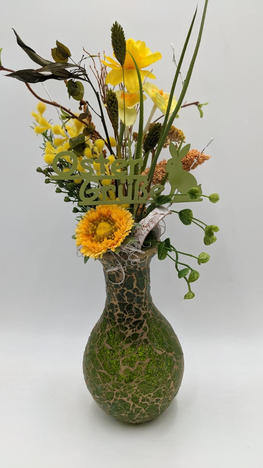 Ostergesteck in bauchiger grüner Vase mit gelben Narzissen, Gerbera, Mimosen, Gräsern und Schriftzug Oster Grüße