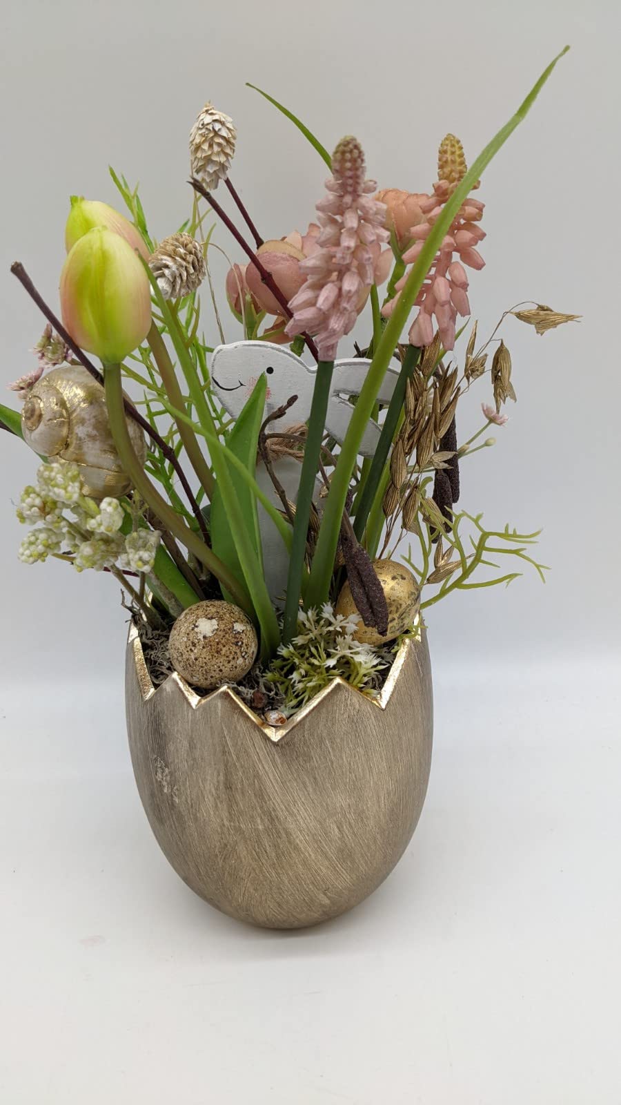 Frühlingsgesteck von vorn: zwei Tulpenknospen, rosa Traubenhyazinthen, Gräser und goldenes Ei im Eierbecher