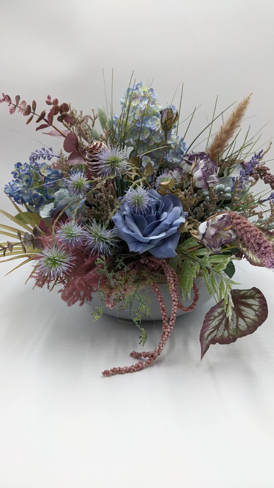 Herbstgesteck mit blauer Rose, blauen Hortensien, Disteln und Gräsern in heller Schale, seitlich fotografiert