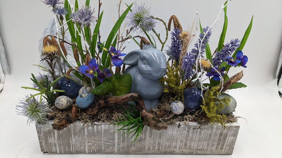 Ostergesteck im grau gekalkten Holzkasten mit graublauem Keramikhasen, blauen Veilchen, Lavendel und gesprenkelten Eiern