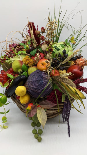 Buntes Erntedank-Gesteck von vorn: grüner Kürbis, Granatapfel, Bananen, Weizenähren und violetter Amaranth im Korb