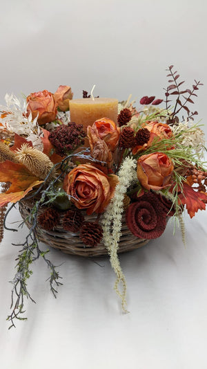 Herbstgesteck im Korb mit ockerfarbener Stumpenkerze, orangen Rosen, Zapfen und hängenden Gräsern