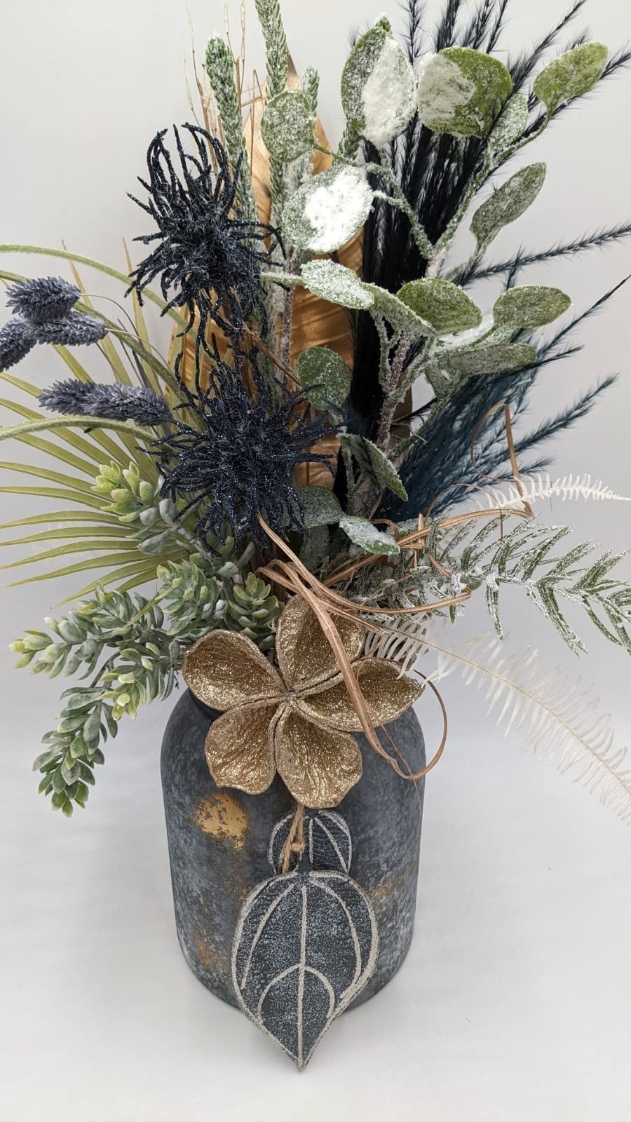 Nahaufnahme des Gestecks: goldene Glitzerblüte mit Bastschleife, blaue Ähren und weiße Farnwedel