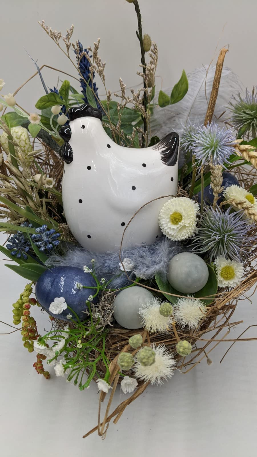 Nahaufnahme des Nests: gepunktetes Keramikhuhn, blaues Ei, hellblaue Federn, Kugeldisteln und weiße Bellis
