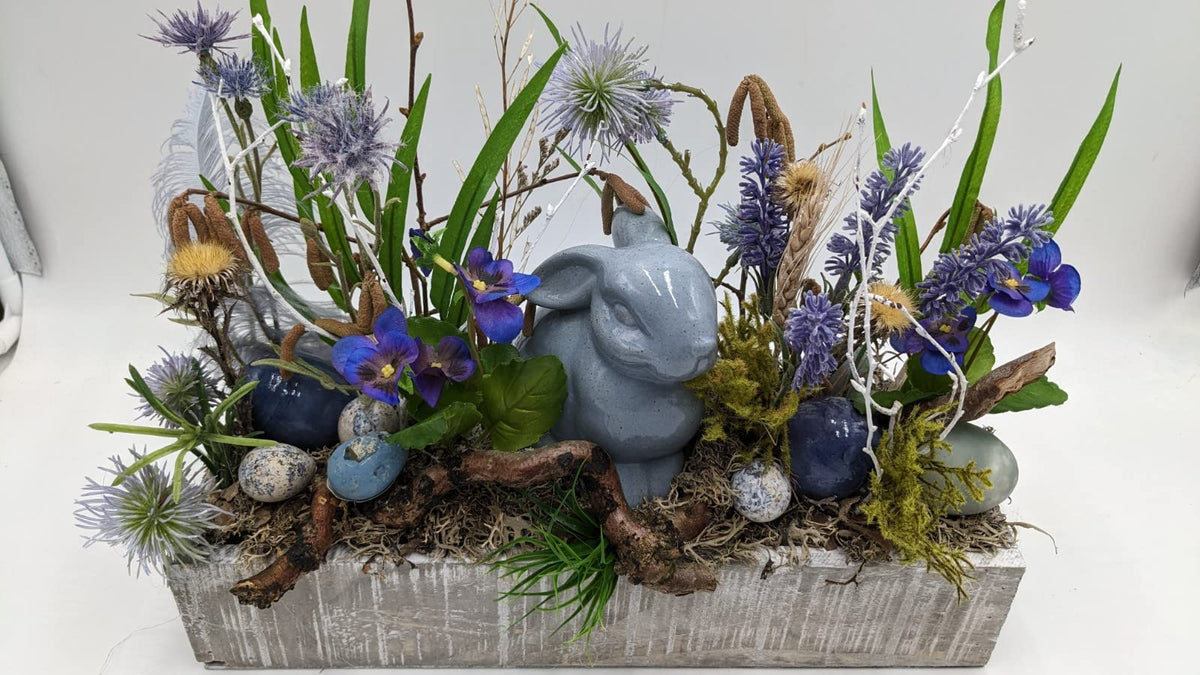 Frühlingsgesteck von der Seite: Hase aus Keramik zwischen Kugeldisteln, Gräsern, Moos, Ast und blauen Ostereiern