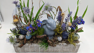 Frühlingsgesteck von der Seite: Hase aus Keramik zwischen Kugeldisteln, Gräsern, Moos, Ast und blauen Ostereiern