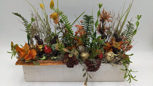Ostergesteck im weißen Holzkasten mit braunen Hasenfiguren, goldenen Eiern, Farn, Gräsern und orangen Blüten