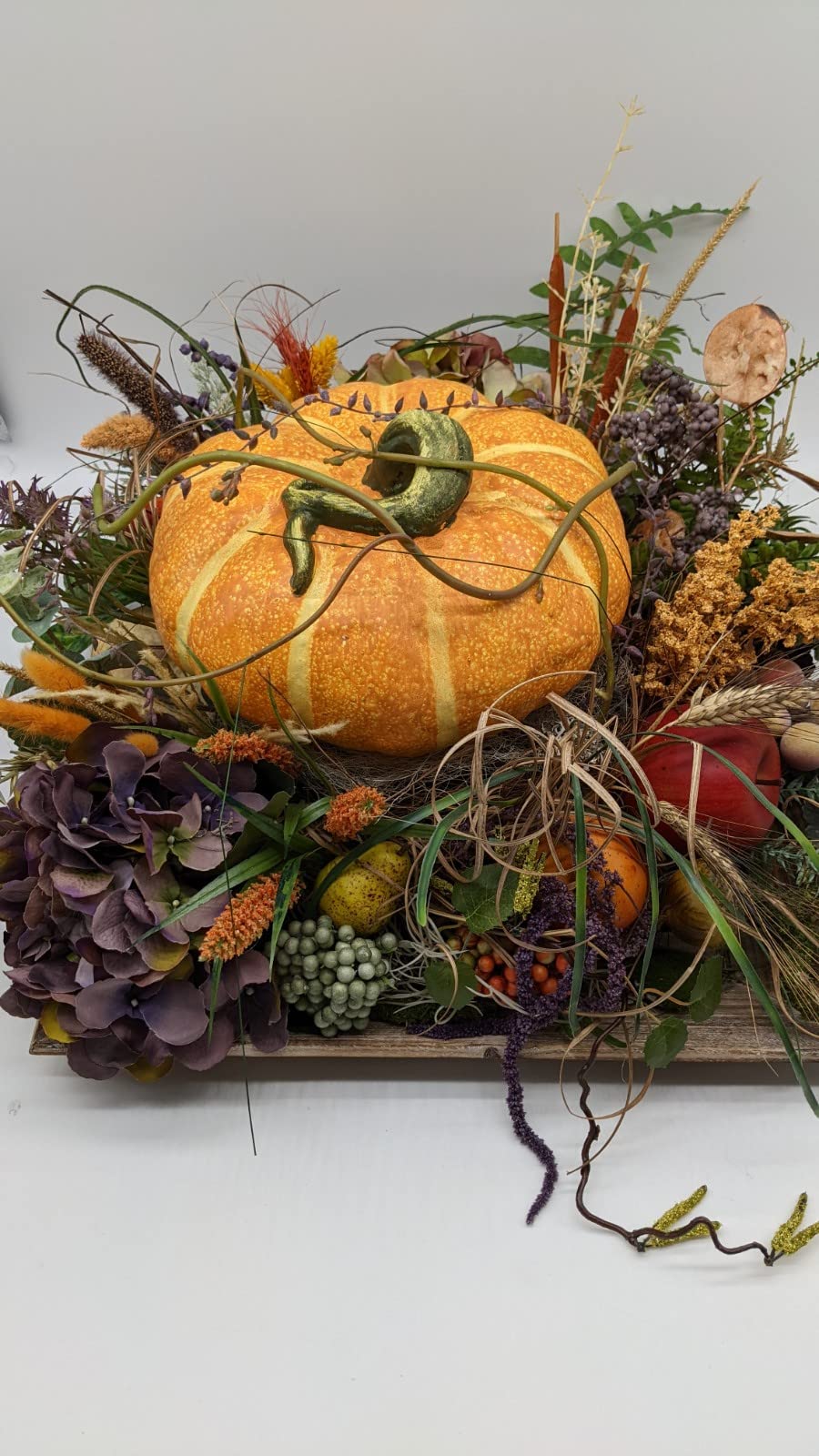 Nahansicht des Herbstgestecks: großer Kürbis mit grünem Stiel, kleine Zierkürbisse, roter Apfel und Bast