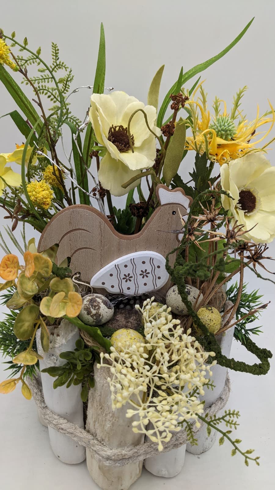 Nahaufnahme des Ostergestecks: Holzhuhn, gesprenkelte Eier, Moos und cremefarbene Beerendolden zwischen gelben Blüten